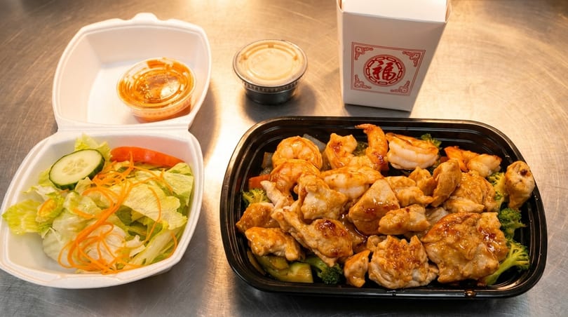 Chinese takeout meal with chicken, shrimp, broccoli, a side salad, and sauces on a metal counter.